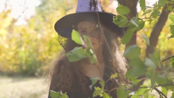Serious redhead caucasian girl dressed in Halloween witch costume standing in the autumn park behind green leaves. Child raising yellow pumpkin and looking at the camera - 映像、動画