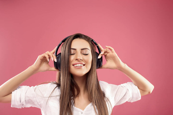 Beautiful young woman in wireless headphones listening to music on pink background. Girl uses wireless earphones - Фото, зображення