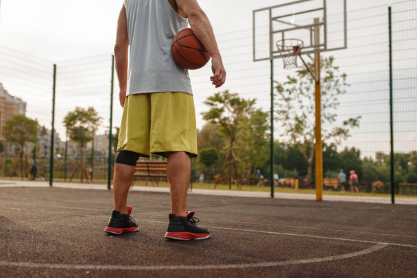 Ανδρική μπασκετμπολίστας με μπάλα στέκεται στο καλάθι στο εξωτερικό γήπεδο, πίσω όψη. Άνδρας αθλητής σε αθλητικά είδη στην προπόνηση streetball, θερινό στάδιο - Φωτογραφία, εικόνα