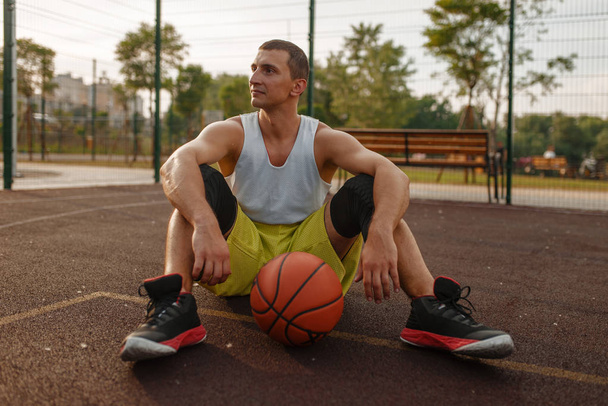 Μπάσκετ παίκτης κάθεται στο έδαφος σε υπαίθριο γήπεδο. Άνδρας αθλητής σε αθλητικά είδη ξεκουράζεται μετά την προπόνηση streetball - Φωτογραφία, εικόνα