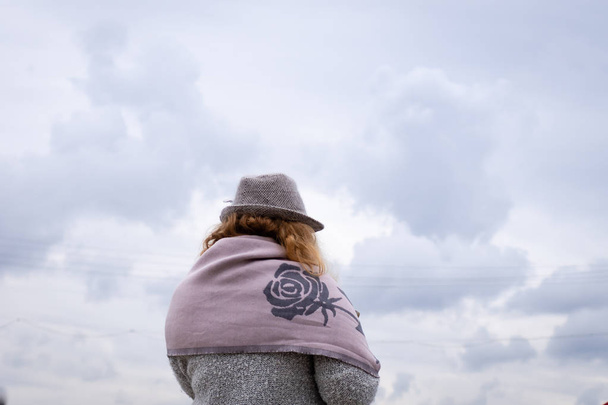 Γυναικεία πλάτη σε ένα ζεστό γκρι παλτό, γκρι καπέλο και ροζ μαντήλι σε φόντο φθινοπωρινού ουρανού με σύννεφα. Οριζόντια κατεύθυνση. Ψυχρή ενότητα. - Φωτογραφία, εικόνα