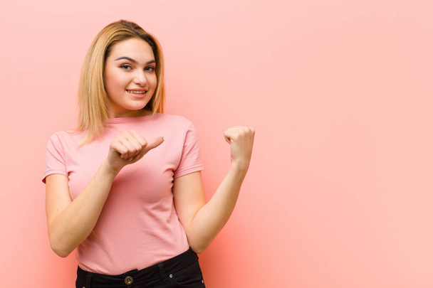 young pretty blonde woman smiling cheerfully and casually pointing to copy space on the side, feeling happy and satisfied against pink flat wall - Fotoğraf, Görsel