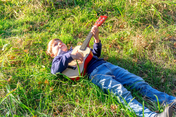 Un niño toca la guitarra tumbado en la hierba verde del parque
. - Foto, Imagen