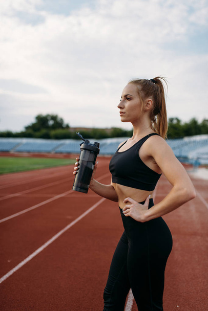 Female runner in sportswear drinks water, training on stadium. Woman doing stretching exercise before running on outdoor arena - Fotografie, Obrázek