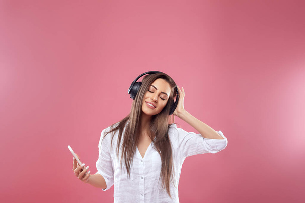 Portrait of a happy young beautiful woman posing isolated over pink wall background listening music with wireless earphones using mobile phone. Girl uses wireless headphones. - Foto, imagen