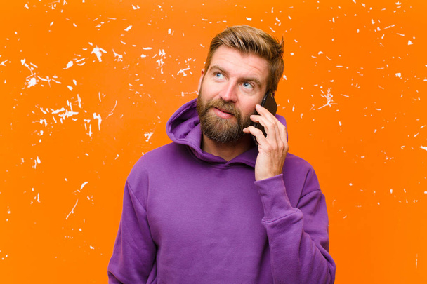 young blonde man with a smart phone wearing a purple hoodie against damaged orange wall - Fotó, kép