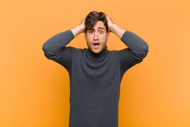 young handsome man looking excited and surprised, open-mouthed with both hands on head, feeling like a lucky winner against orange background - Foto, Imagen
