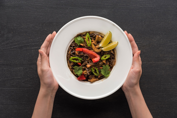cropped view of woman holding plate with tasty spicy thai noodles on wooden grey surface - Фото, изображение