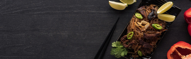 top view of meat thai noodles near chopsticks, lime and paprika on wooden grey surface, panoramic shot - Photo, Image