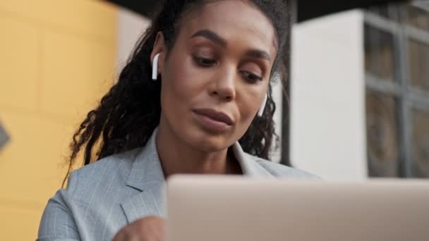 Close up view of serious young african business woman listening radio with earpods and waiting for someone while working on laptop in cafe - Footage, Video