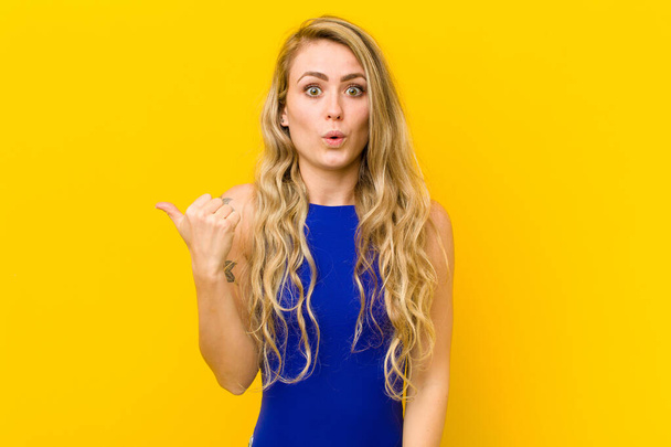 young blonde woman looking astonished in disbelief, pointing at object on the side and saying wow, unbelievable against yellow wall - Photo, Image