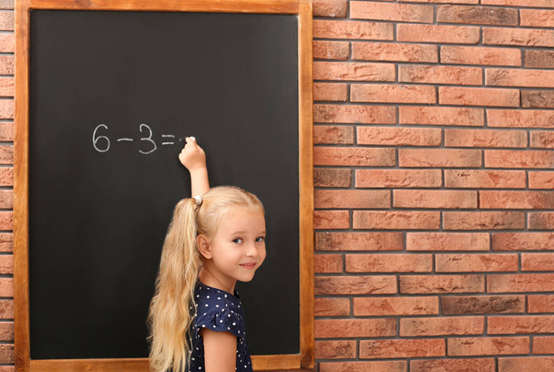 Cute little left-handed girl doing sums on chalkboard near brick wall - Foto, Imagen