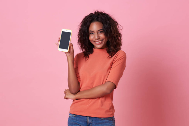 Portrait de heureuse jeune femme africaine montrant à l'écran vierge téléphone portable isolé sur fond rose
. - Photo, image
