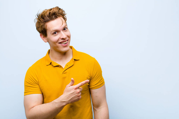 young red head man looking excited and surprised pointing to the side and upwards to copy space against blue wall - Foto, immagini