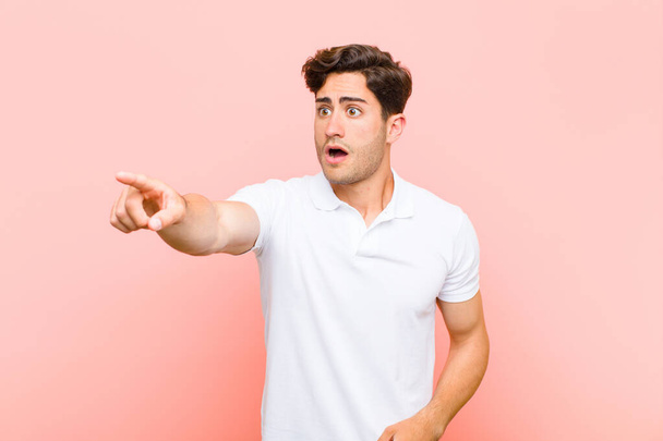 young handsome man feeling shocked and surprised, pointing and looking upwards in awe with amazed, open-mouthed look against pink background - Foto, Bild