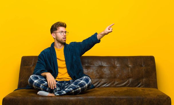 young man wearing pajamas feeling shocked and surprised, pointing and looking upwards in awe with amazed, open-mouthed look . sitting on a sofa - Fotoğraf, Görsel