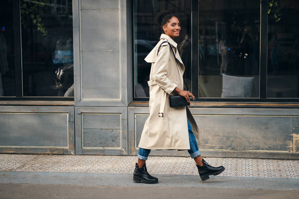 Side view of smiling casual African American girl in trench coat happily looking in camera walking down the street - Photo, Image