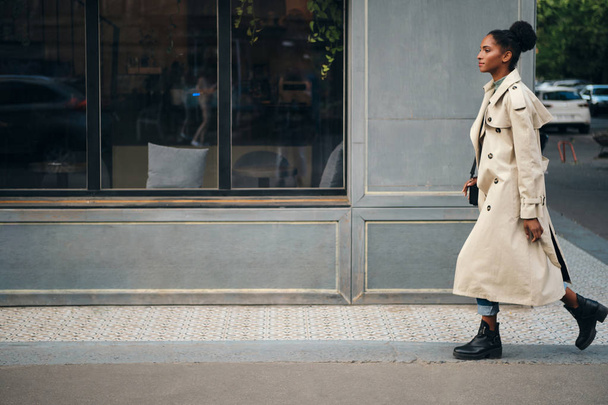 Side view of attractive casual African American girl in trench coat dreamily walking down the street - Foto, Bild
