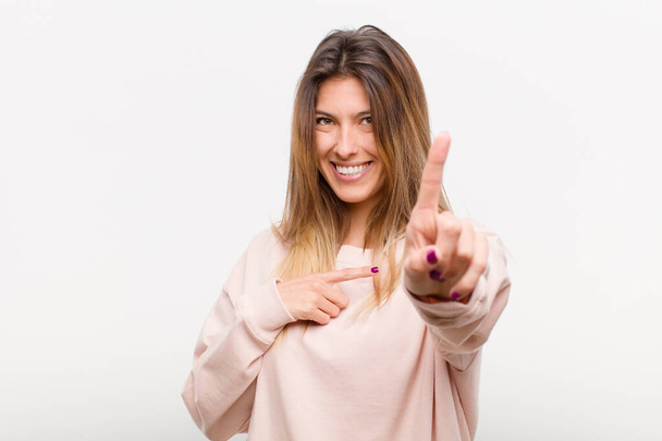 young pretty woman smiling proudly and confidently making number one pose triumphantly, feeling like a leader against white background - Фото, изображение