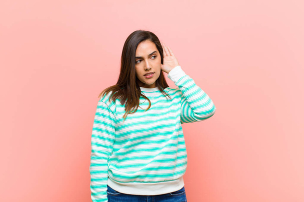 young pretty hispanic woman looking serious and curious, listening, trying to hear a secret conversation or gossip, eavesdropping against pink wall - Photo, Image