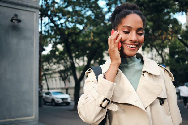Aantrekkelijk lachend Afro-Amerikaans meisje in stijlvolle trench jas vrolijk praten op mobiele telefoon in de stad straat - Foto, afbeelding