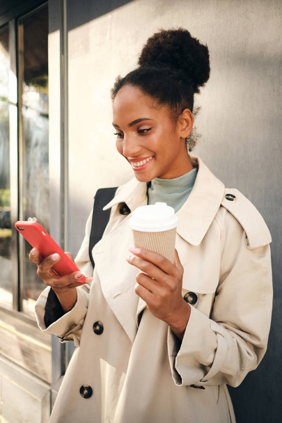 Güzel gülümseyen Afro-Amerikan kız şık trençkotlu ve kahveli açık havada mutlu bir şekilde cep telefonu kullanıyor. - Fotoğraf, Görsel