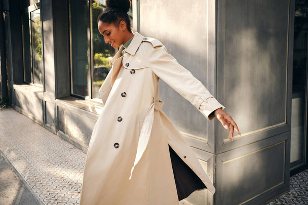 Side view of attractive casual African American girl in stylish trench coat happily whirling on city street - Фото, зображення