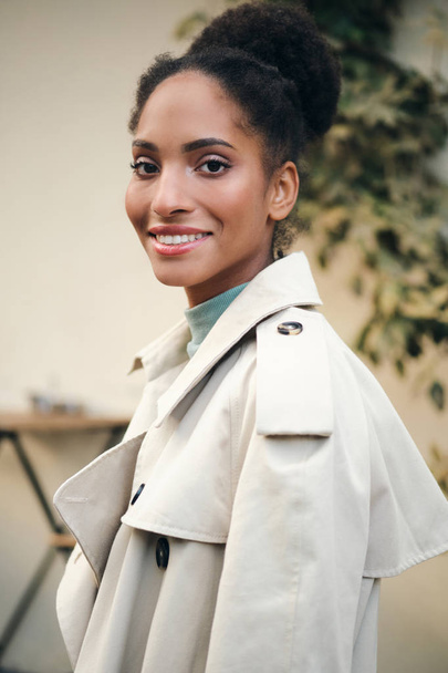 Retrato de una chica afroamericana bastante sonriente con un elegante abrigo de trinchera mirando felizmente en la cámara en la calle
 - Foto, imagen