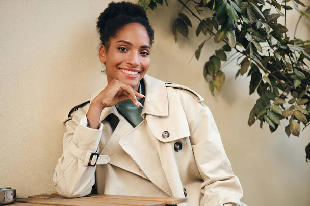 Cheerful African American girl in stylish trench coat joyfully looking in camera in cafe on street - 写真・画像