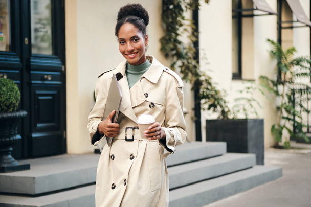 Pretty casual African American student girl in stylish trench coat with laptop and coffee to go joyfully looking in camera on city street - Φωτογραφία, εικόνα