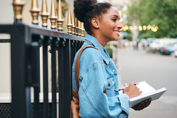 Side view of beautiful smiling casual African American student girl in denim jacket with notepad happily studying on city street - Φωτογραφία, εικόνα
