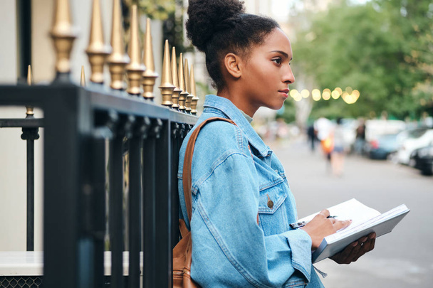 Vista lateral da menina estudante afro-americana casual em jaqueta jeans com bloco de notas estudando pensativamente na rua da cidade
 - Foto, Imagem