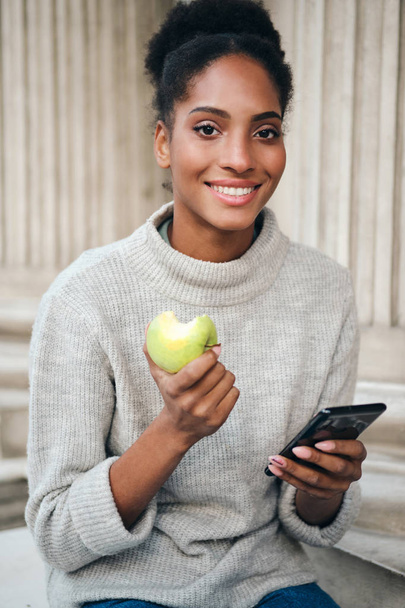 Attractive smiling African American girl in sweater with cellphone eating apple happily looking in camera on city street - Photo, Image