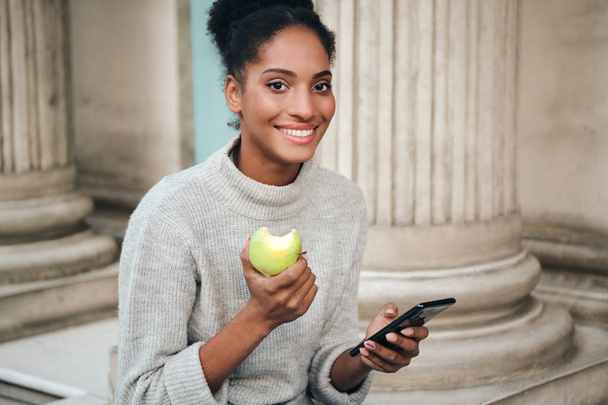Beautiful smiling African American girl in sweater with cellphone eating apple joyfully looking in camera outdoor - Φωτογραφία, εικόνα