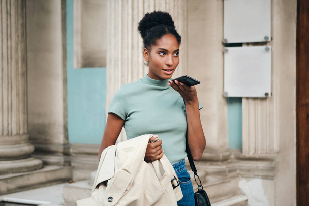 Belle fille afro-américaine avec trench coat à la main parlant avec réflexion sur le téléphone portable sur la rue de la ville
 - Photo, image
