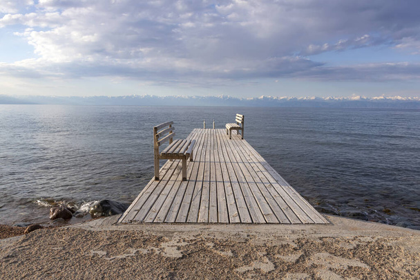 Molo s lavičkami na jezeře Issyk-Kul s pohořím se zasněženými vrcholky na obzoru a zamračenou oblohou - Fotografie, Obrázek