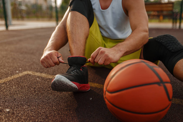 Basketball player sitting on the ground and tying laces on outdoor court. Male athlete in sportswear resting after streetball training - Foto, afbeelding