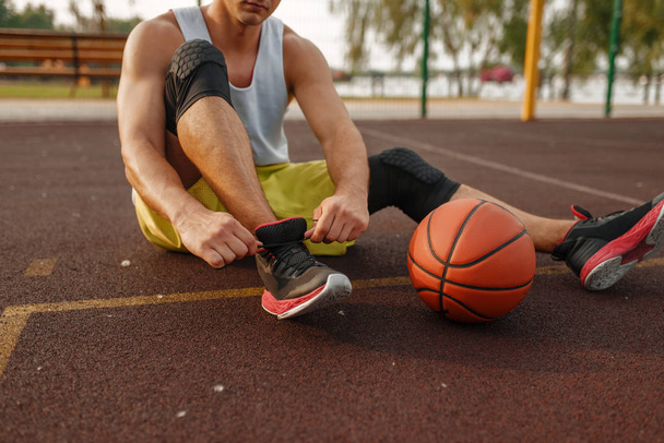 バスケットボール選手は地面に座り、屋外コートでレースを結びます。スポーツウェアの男性選手は、ストリートボールのトレーニング後に休息 - 写真・画像