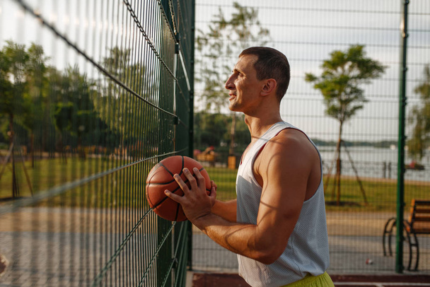 Jugador de baloncesto masculino con pelota de pie en la valla de malla en la cancha al aire libre, vista lateral. Atleta masculino en ropa deportiva en entrenamiento de streetball, estadio de verano
 - Foto, Imagen