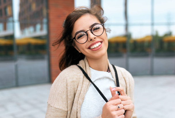 Outdoors portrait of pretty young woman student smiling broadly with toothy smile, with backpack on the back, wearing transparent eyeglasses looking to the camera, posing on the city street. - Fotografie, Obrázek