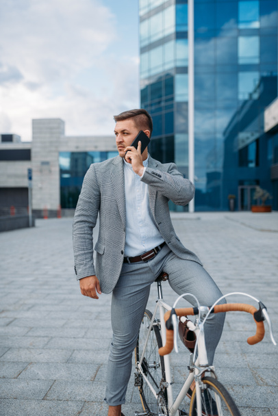 Businessman in suit talking by phone on the bicycle in downtown. Business person riding on eco transport on city street - Fotó, kép