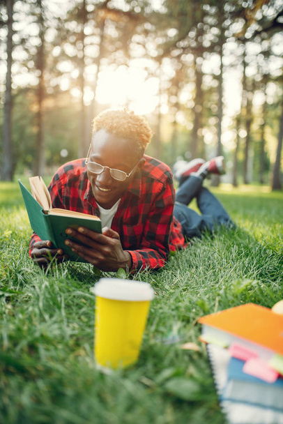 Африканский студент в наушниках лежит на траве и читает книгу в летнем парке
.  - Фото, изображение