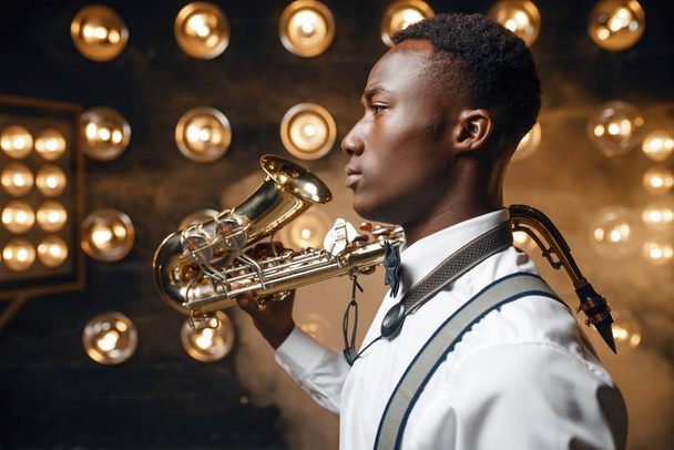 Afrikaanse mannelijke jazz performer poseert met saxofoon op het podium met schijnwerpers.  - Foto, afbeelding