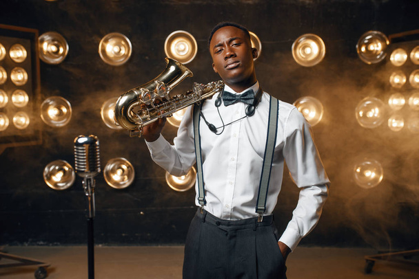 Afrikaanse mannelijke jazz performer poseert met saxofoon op het podium met schijnwerpers.  - Foto, afbeelding