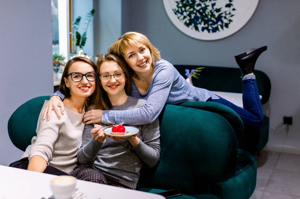 Women friends sitting on the sofa in cafe, looking at camera, one girl holds plate with delicious red dessert - Zdjęcie, obraz