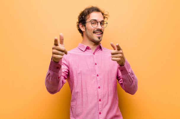 young handsome man smiling with a positive, successful, happy attitude pointing to the camera, making gun sign with hands against orange wall - Φωτογραφία, εικόνα