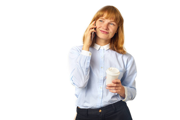 young redheaded woman with freckles and bang talking on phone and smiling while holding cup of coffee in hand  - Фото, зображення