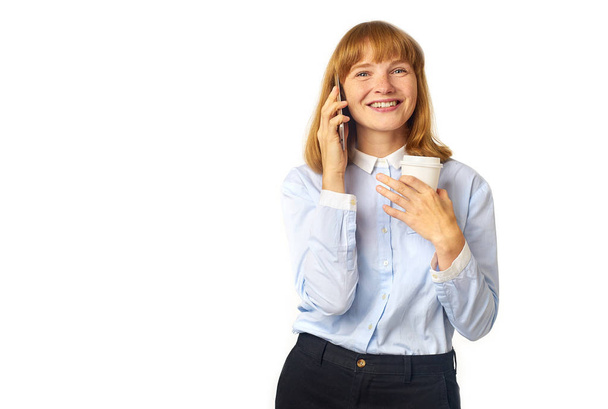 young redheaded woman with freckles and bang talking on phone and smiling while holding cup of coffee in hand  - Photo, image