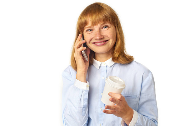 young redheaded woman with freckles and bang talking on phone and smiling while holding cup of coffee in hand  - Photo, Image
