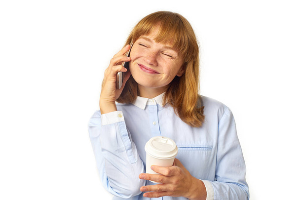 happy female model with ginger hair and freckles dressed in blue shirt talking on smartphone and holding coffee cup while posing with closed arms isolated on white background - Photo, image